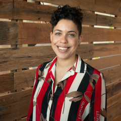 Photo of Jess smiling wearing a striped red and black shirt standing in front of wood panelling