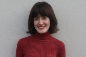 Róisín Ní Riain headshot. In the image she is wearing a red roll neck jumper and is standing against a white wall.