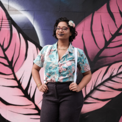 Photo of Chandrika wearing a blue shirt and suspenders standing in front of a wall with a pink leaf mural 