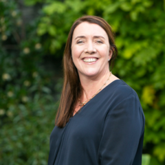 Photo of Aine Denn wearing a navy top, smiling and standing in front of a hedgerow 