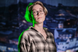 Kel Menton headshot - Kel is wearing a plaid shirt and glasses