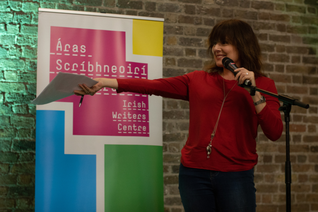 Performer Tara Flynn stands in front of a microphone and holds her arm out