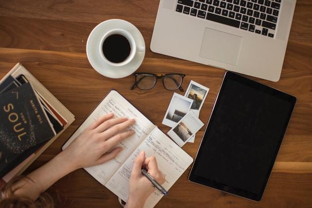 Photo of a desk with someone writing in a notebook, coffee, laptop and books